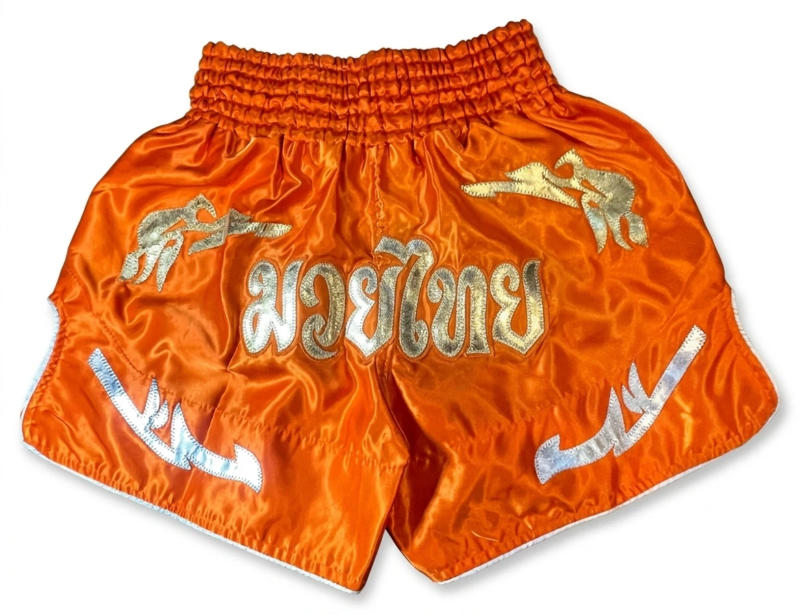 Back view of vibrant orange satin Muay Thai shorts with elastic waist and traditional gold Thai script appliqués.