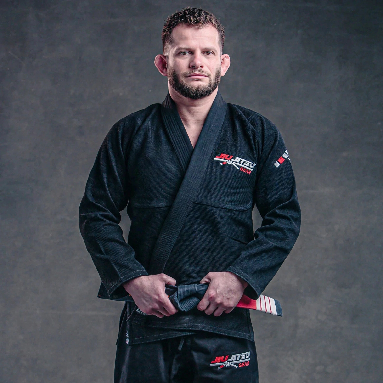 Male BJJ black belt athlete wearing black Brazilian Jiu Jitsu Gi set - JIU JITSU GEAR pearl weave gi jacket and ripstop pants with embroidered red white logo on chest and thigh